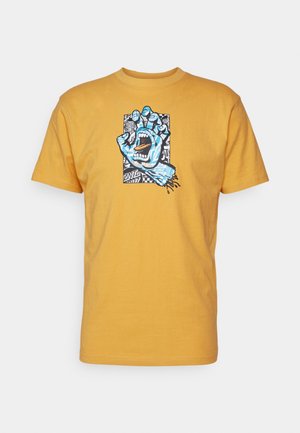T-shirt en coton jaune avec un graphique bleu représentant une main avec une bouche ouverte, sur un fond à motifs noir et blanc.