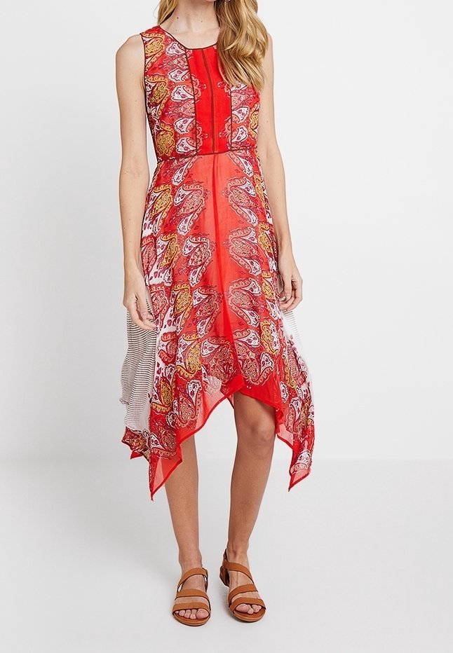 Femme portant une robe asymétrique rouge sans manches à motif cachemire et des sandales marron sur fond blanc.