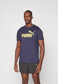 T-shirt à manches courtes bleu marine en coton, présentant un grand logo "PUMA" jaune et une silhouette de chat sur le devant. Assortie à un short noir.