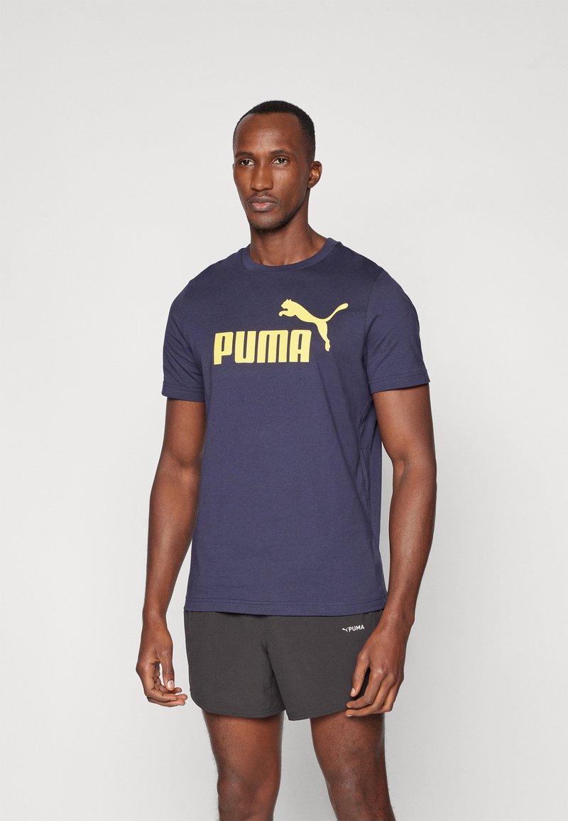 T-shirt à manches courtes bleu marine en coton, présentant un grand logo "PUMA" jaune et une silhouette de chat sur le devant. Assortie à un short noir.
