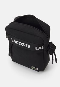 Black fabric shoulder bag with a zippered closure, featuring a white "LACOSTE" logo strap and a green crocodile logo. Textured surface.