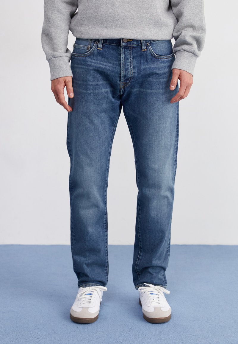 Lichtblauwe denim jeans met een rechte pasvorm, vijf zakken, subtiele vervaging en een knoopsluiting. Gdragen met witte sneakers.