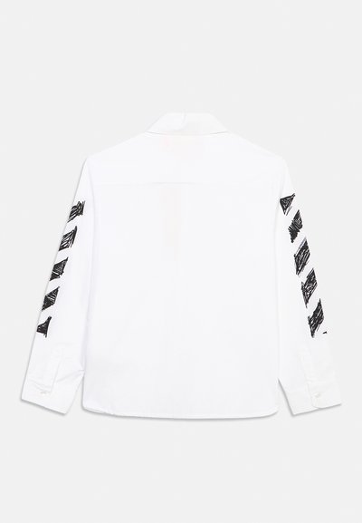 Λευκό πουκάμισο με μακριά μανίκια, εμφανιζόμενο από πίσω, με μαύρα αφηρημένα σχεδιαγράμματα που τρέχουν κατά μήκος και των δύο μανικιών.