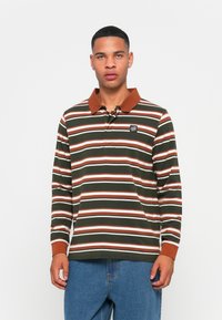 Man in een groen, bruin en wit gestreepte lange mouwen polo met een bruine kraag en manchetten, gecombineerd met blauwe jeans, staande tegen een witte achtergrond.
