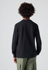 Juodas ilgomis rankovėmis marškinėliai pagaminti iš medvilnės, turintys apvalų iškirpimą, matomi iš nugaros, derinami su alyvuogių spalvos cargo kelnėmis.