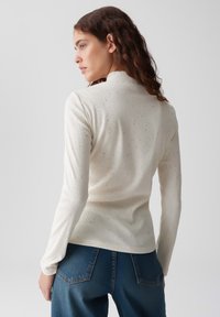 Langärmliges, geripptes Turtleneck-Oberteil in Creme mit dezenten Sprenkeln. Weiche Textur, taillierte Passform und hoher Kragen. Getragen mit blauen Denim-Jeans.