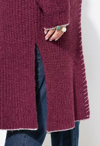 Bordový pletený kardigan má texturovanou tkaninu, boční rozparky a bílé švy na lemech a okrajích rukávů.