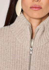 Frau, die einen beigen, gerippten Strickpullover mit Reißverschluss und großen ovalen beigen Ohrringen trägt, wobei der untere Gesichtsteil und der Halsbereich zu sehen sind.