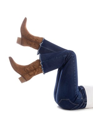 Botas de vaquero de ante marrón con puntas afiladas, que presentan costuras decorativas, combinadas con jeans de campana azul oscuro con dobladillos deshilachados.