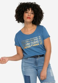 Camiseta de algodón azul con mangas cortas enrolladas y escote redondeado, que presenta el texto metálico dorado "UNDER THINK IT" en un diseño en capas.