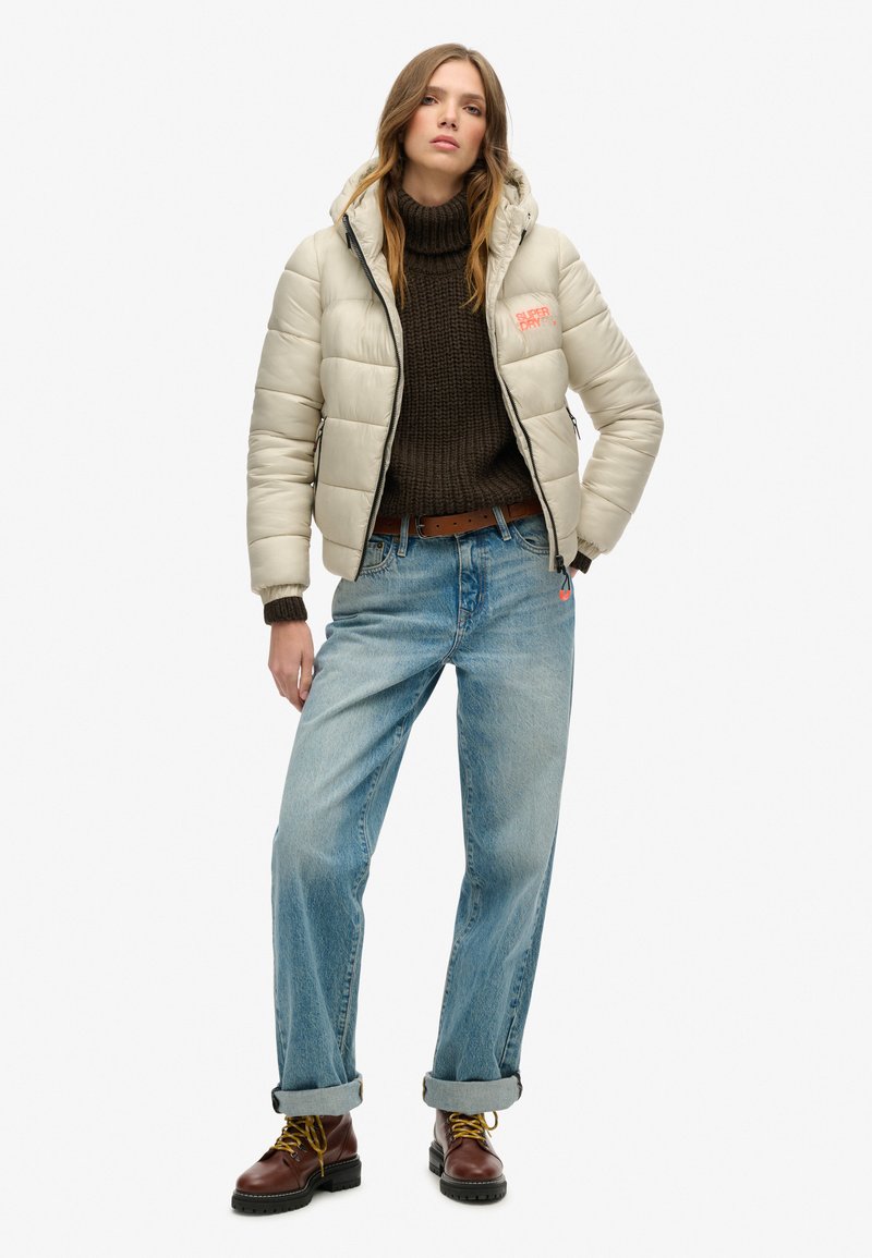 Beige Pufferjacke mit Kapuze, strukturiertem Stoff, mit Reißverschlusstaschen, kombiniert mit einem dunklen Strickpullover und hellblauen, locker sitzenden Jeans, braunen Schnürstiefeln.