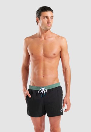 Hemdloser Mann trägt schwarze Badeshorts mit grünem Bund und weißer Kordel, steht mit einer Hand in der Tasche und schaut nach rechts.