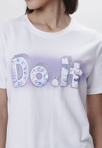 Witte T-shirt met een verloop in paarse glans. "Doe het" versierd met iriserende pailletten en edelsteen-achtige accenten voor een textuur effect.