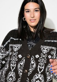 Brun stickad cardigan med vita geometriska mönster, med V-ringning, tre knappar och ribbad mudd. Texturerad ulltyg.
