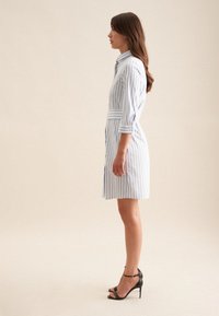 Kvinde stående sideværts iført en knælængde, blå og hvide lodrette stribet kjole med opkørte ærmer og sorte højhælede sandaler.