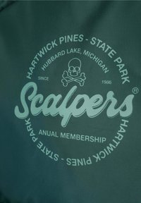 La tela verde presenta un logo circular con el texto "Scalpers", un diseño de calavera y huesos cruzados, y los detalles del nombre del parque en una impresión verde claro.