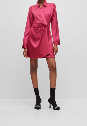 Frau trägt ein rosa Satin-Wickelkleid mit langen Ärmeln, kombiniert mit schwarzen Sockboots mit Absatz und Strasssteindetails an den Zehen.