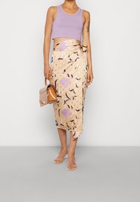 Débardeur court violet clair assorti à une jupe portefeuille mi-longue à motif floral beige, avec des accents violets et bleus. Sandales à talons transparents.