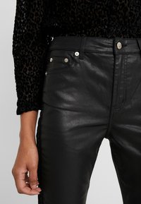 Zwarte broek van nep leer met een glanzende afwerking, voorzien van zilverkleurige hardware en zijstikseldetails. Draagt een gestructureerde zwarte top.