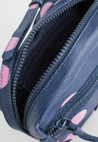 Zila sintētiskā soma ar rozā apļveida akcentiem, ar dubultiem zīmuļiem un teksturētām virsmām, kurā redzami iekšējie nodalījumi un siksnas.