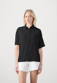 Svart kortermet skjorte med knappestolpe og krage samt brystlomme, laget av et glattekstilt stoff. Kombinert med hvite denimshorts med slitte kanter.