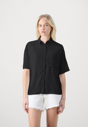 Black short-sleeve button-up shirt with a collar and a chest pocket, made of a smooth fabric. Paired with white denim shorts featuring frayed hems.