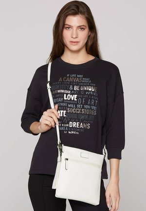 White crossbody bag with a textured surface, featuring a zippered pocket and an adjustable strap. Model wears a black graphic sweatshirt.