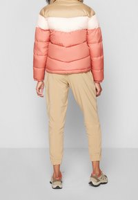 Kobieta ubrana w pikowaną kurtkę typu color-block w kolorach beżowym, brzoskwiniowym i koralowym, beżowe spodnie dresowe oraz beżowe sneakersy z koralowymi akcentami, stojąca tyłem.