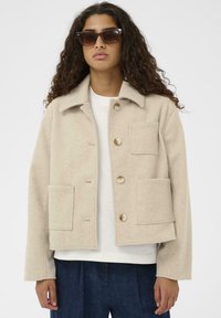Veste en laine beige mélangée avec un col, quatre poches avant et des boutons dorés. Coupe décontractée et tissu texturé.