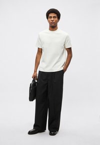 Jasnokremowa bluzka z krótkim rękawem w połączeniu z szerokimi czarnymi spodniami. Modelka trzyma czarną skórzaną torbę, stojąc na neutralnym tle.