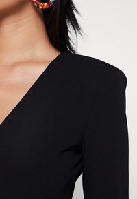 Top nero a V con una trama liscia e spalle strutturate. Orecchini colorati e con perline aggiungono un tocco di dettaglio. Focus sulla scollatura e sul profilo delle maniche.