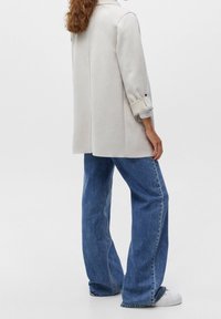 Femme aux cheveux bouclés portant un blazer beige clair, un jean large bleu et des baskets blanches, debout devant un fond uni.