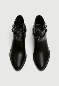 Botines de cuero negro hasta el tobillo con punteras puntiagudas, que incluyen una correa envolvente con tachuelas y detalles de hebilla metálica. Textura suave en general.