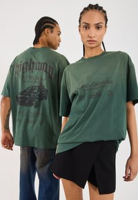 To personer bærer løstsiddende, olivengrønne t-shirts med grafik, der viser tekst og bildesigns. Kvinden kombinerer sin t-shirt med en sort nederdel.