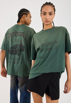 Två individer bär löst sittande, olivgröna grafiska t-shirts med text och bild på bilar. Kvinnan matchar sin med en svart kjol.