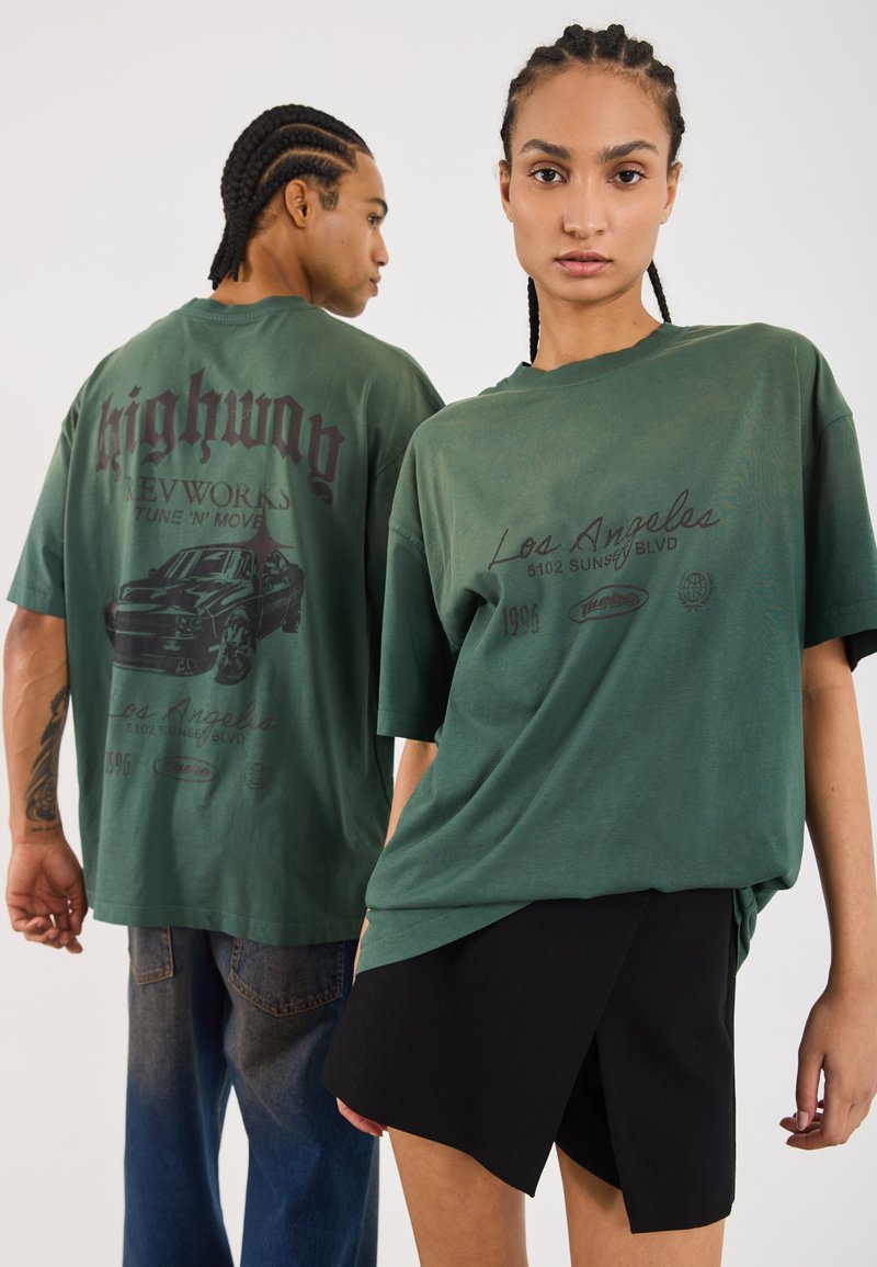 To personer bærer løstsiddende, olivengrønne t-shirts med grafik, der viser tekst og bildesigns. Kvinden kombinerer sin t-shirt med en sort nederdel.