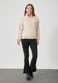 Beige fleece trui met een halve rits en lange mouwen, gecombineerd met zwarte uitlopende broek en zwarte enkellaarsjes. Gladde textuur.
