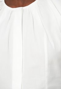 Blusa branca com tecido suave e acabamento texturizado, detalhes plissados ao longo da linha do decote e colarinho largo e arredondado.