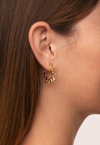 Nahaufnahme eines Frauenohrs mit glattem braunem Haar, das einen strukturierten goldenen Creolenohrring in Form unregelmäßiger Spitzen trägt.