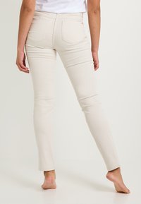 Jeans skinny beige con una textura suave, cintura de media altura, dos bolsillos traseros y una silueta ajustada, mostrados desde la vista trasera.