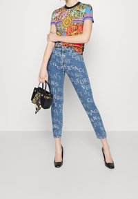 T-shirt grafico con disegni ornamentali colorati abbinato a jeans in denim blu impreziositi con la scritta "Versace" in un motivo ripetuto. Borsa nera e tacchi.