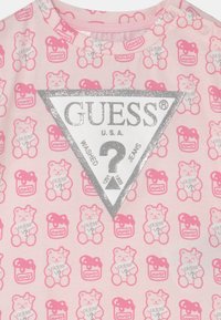 Svetle růžové tričko s potiskem medvídka a sklenice s medem, s metalicky stříbrným trojúhelníkovým výšivkou s logem a textem "GUESS".