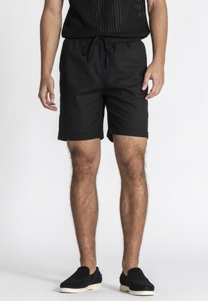 Mann trägt schwarze Shorts mit Kordelzug, schwarzes ärmelloses Stricktop und schwarze Slip-On-Schuhe mit weißen Sohlen, steht vor einem weißen Hintergrund.