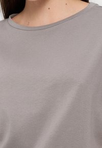 Κοντινό πλάνο του λαιμού και του πάνω μέρους του στήθους ενός ατόμου που φοράει ένα απλό ανοιχτό γκρι μπλουζάκι με στρογγυλή λαιμόκοψη.