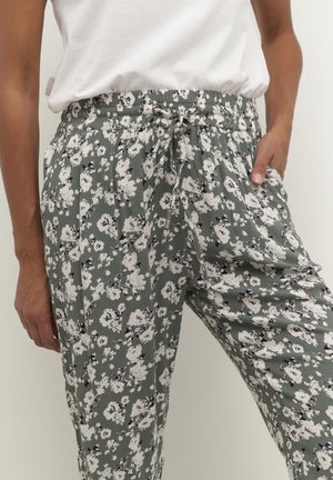 Vrouw draagt een broek met een groen trekkoord en bloemenprint, en een wit mouwloos topje ingestopt, met handen in de zakken.