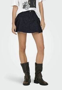 Denim-minikjol med fransar i kanten, mörkblå färg, med sidfickor och knappstängning fram, kombinerad med svarta anklarstövlar.