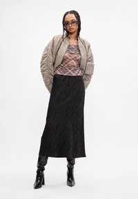 Casaco bomber em tecido cinza claro, blusa xadrez em cores suaves, saia longa preta com padrão abstrato e botas pretas brilhantes até ao joelho.