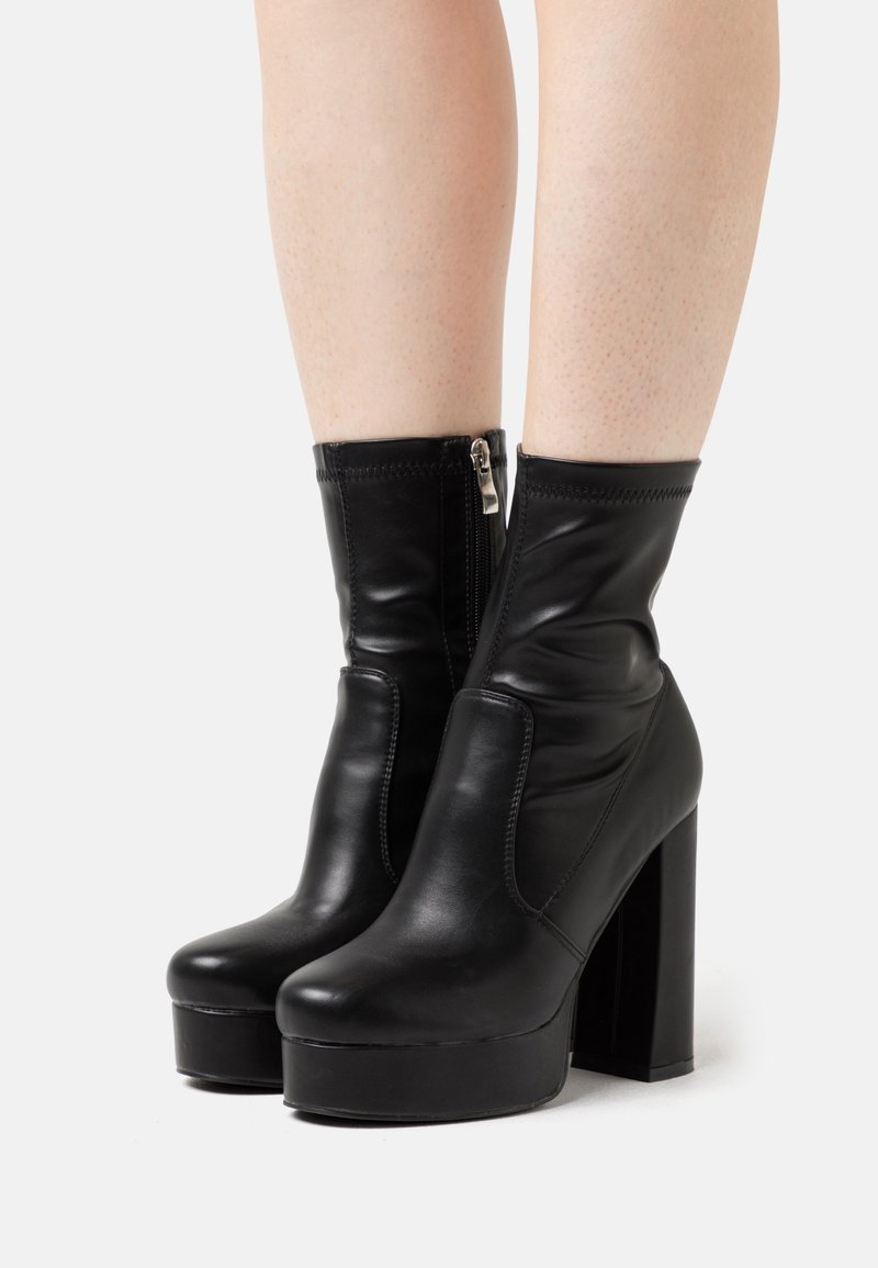 Bottes noires en cuir à plateforme, à talons hauts épais et fermeture éclair latérale, portées sur des jambes nues sur fond blanc.