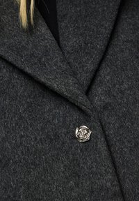 Σκούρο γκρι μάλλινο παλτό με αυστηρό γιακά, διαθέτοντας ένα ασημένιο κουμπί σε σχήμα τριαντάφυλλου ως ξεχωριστή λεπτομέρεια. Μαλακή υφή και ραμμένο σχέδιο.
