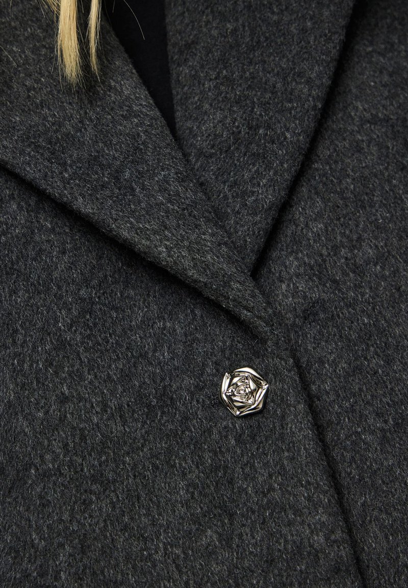 Mörkgrå ullrock med notch-krage, med en silverfärgad ros-formad knapp som en särskild detalj. Mjukt material och skräddarsydd design.